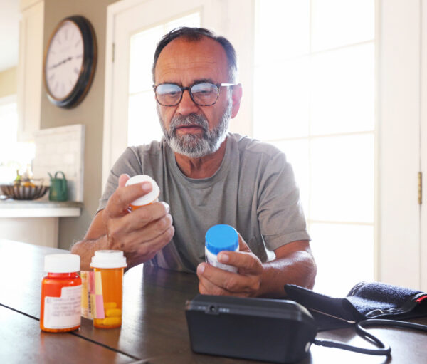 A Latino senior citizen sits at his dining room table and sorts through various prescription medications as sunlight filters in through the window behind him bathing the room with a soft glow of light. A blood pressure machine rests on the table in front of him.
