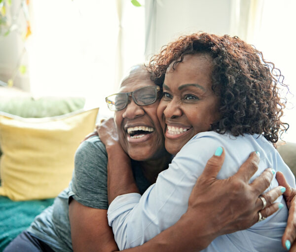 Waist-up view of Black women in early 50s and late 60s sitting on sofa in family home, cheek to cheek and grinning as they embrace.