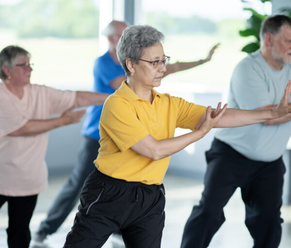 A small group of seniors are seen practicing Tai Chi in an indoor studio. They are each dressed comfortably and are focused on their movements and proper form as they practice the Martial Art.